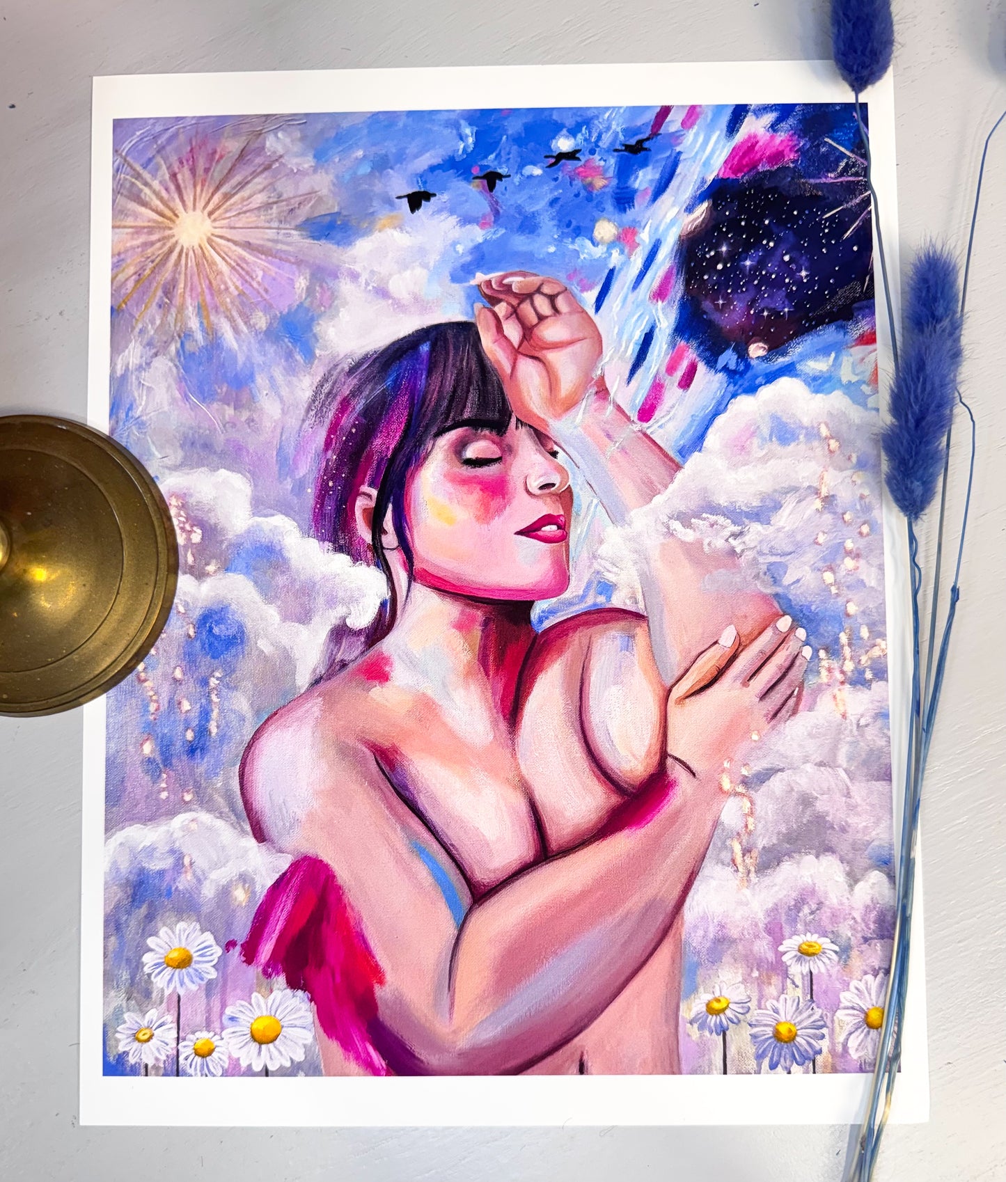 A picture of an art print featuring a nude woman with her eyes closed in the clouds surrounded by flowers, birds, a sun, stars and a galaxy.