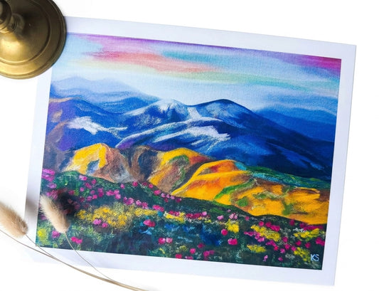 A print featuring a landscape with blue and yellow mountains and a field of magenta pink flowers 