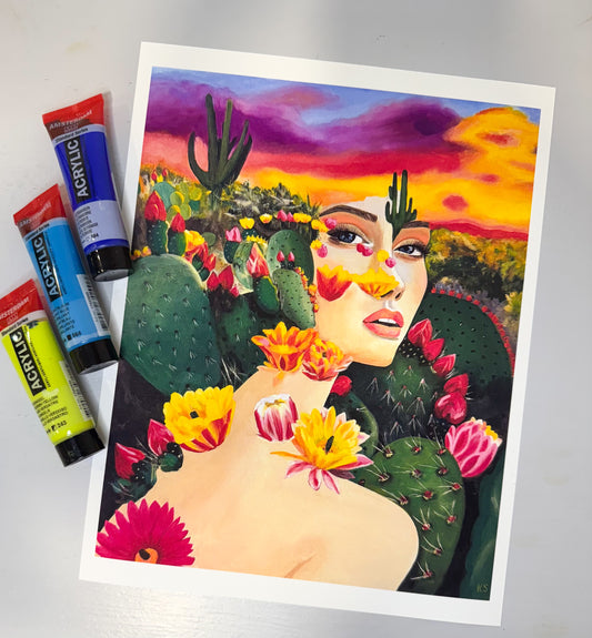 Colorful art print of a woman with cacti and flowers, next to acrylic paint tubes on a grey surface.