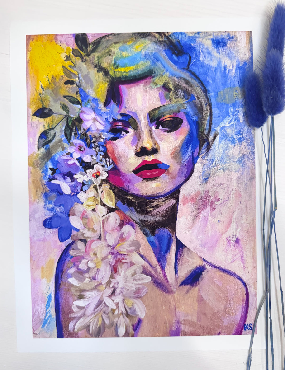 A colorful portrait painting of a woman with blue, purple, pink, and yellow tones, surrounded by flowers, displayed on a white surface with blue decorative grass stems on the right.