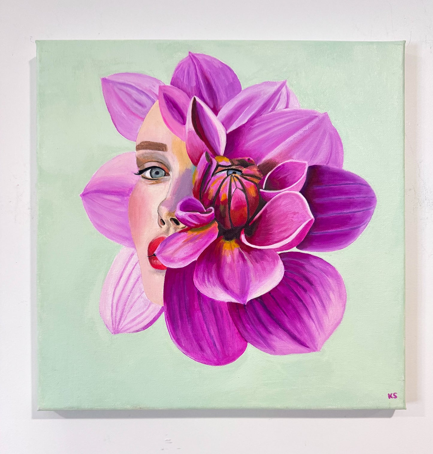 A square canvas painting featuring a stylized pink and purple flower with a woman's face emerging from behind it, set against a pale mint green background. The canvas is shown hanging on a white wall.