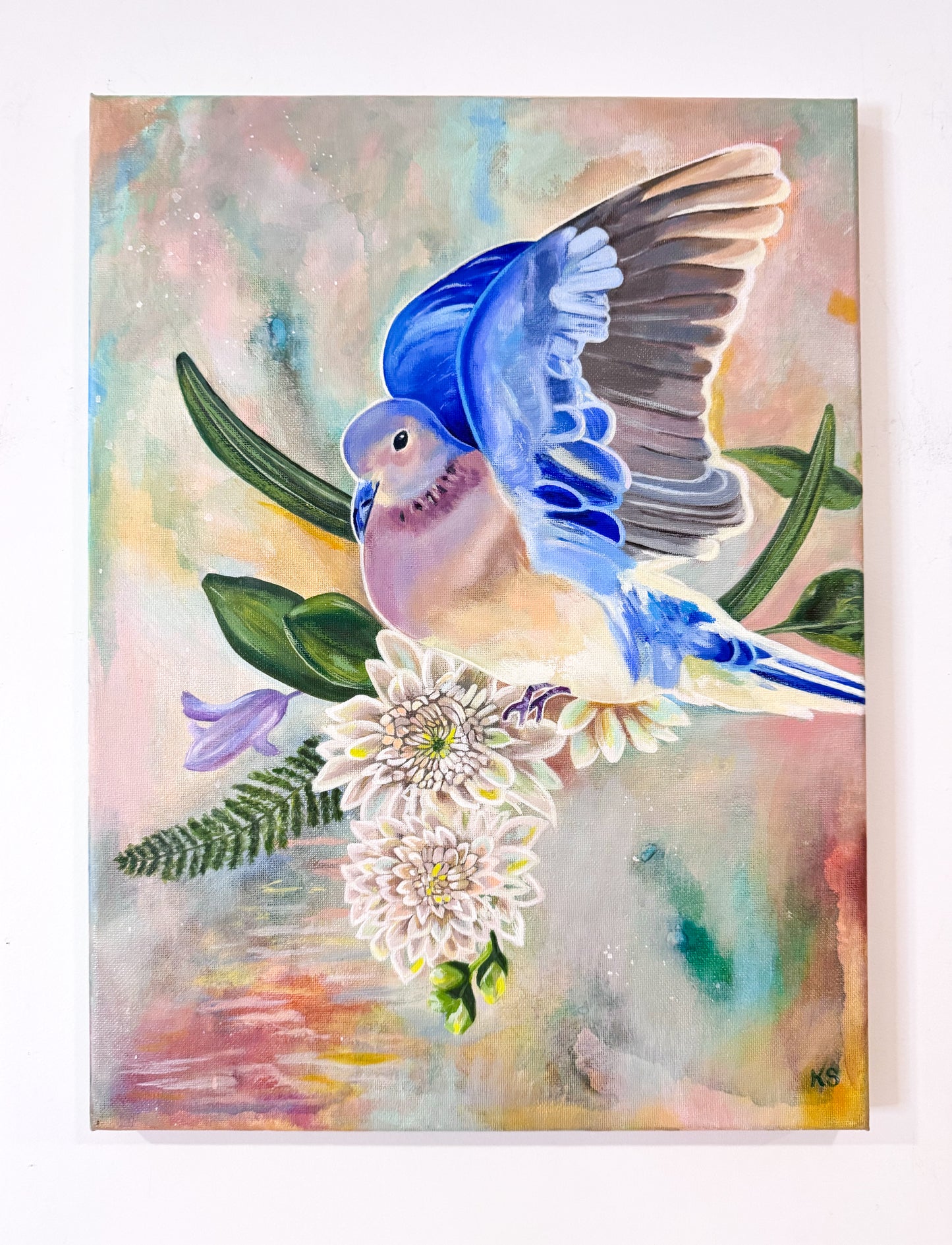 A colorful watercolor-style painting of a blue bird with spread wings perched on white flowers and green leaves, set against a soft pastel background. The artwork is shown hanging on a white wall.