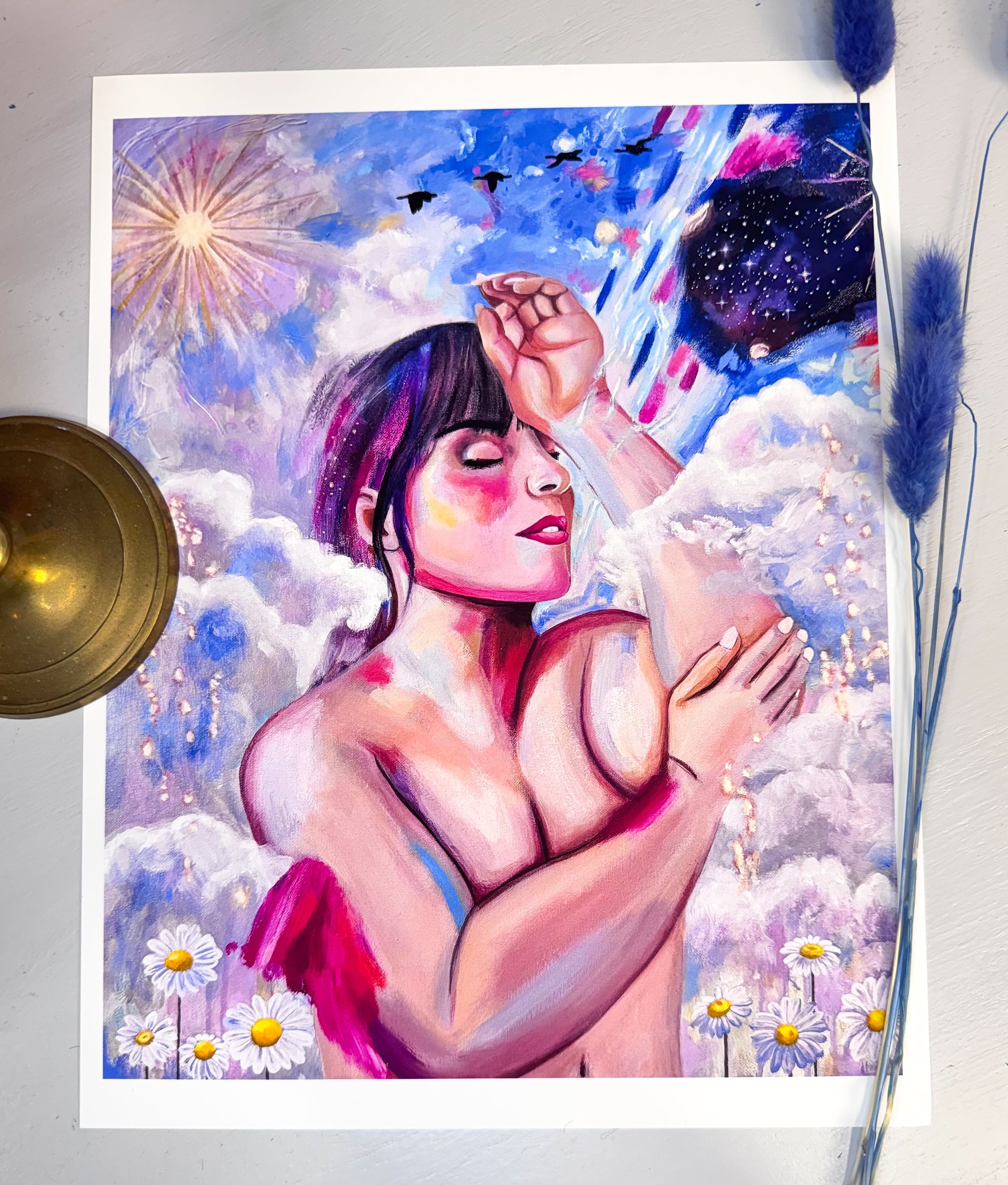 A picture of an art print featuring a nude woman with her eyes closed in the clouds surrounded by flowers, birds, a sun, stars and a galaxy.