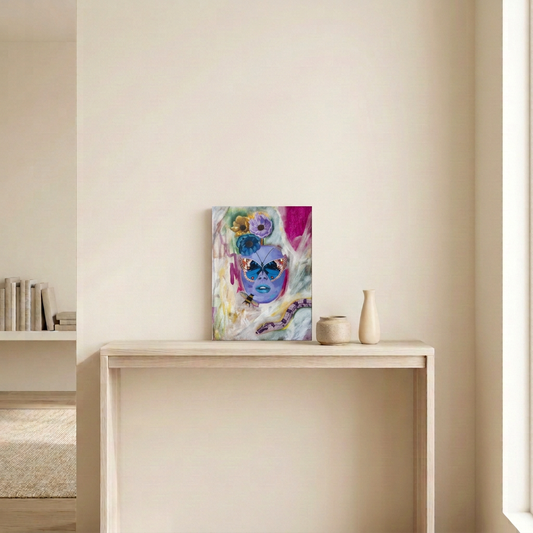 Ethereal Creature, a surreal painting of a purple face with a butterfly, flowers, bee and snake, displayed unframed on a pale wood console in a neutral room.